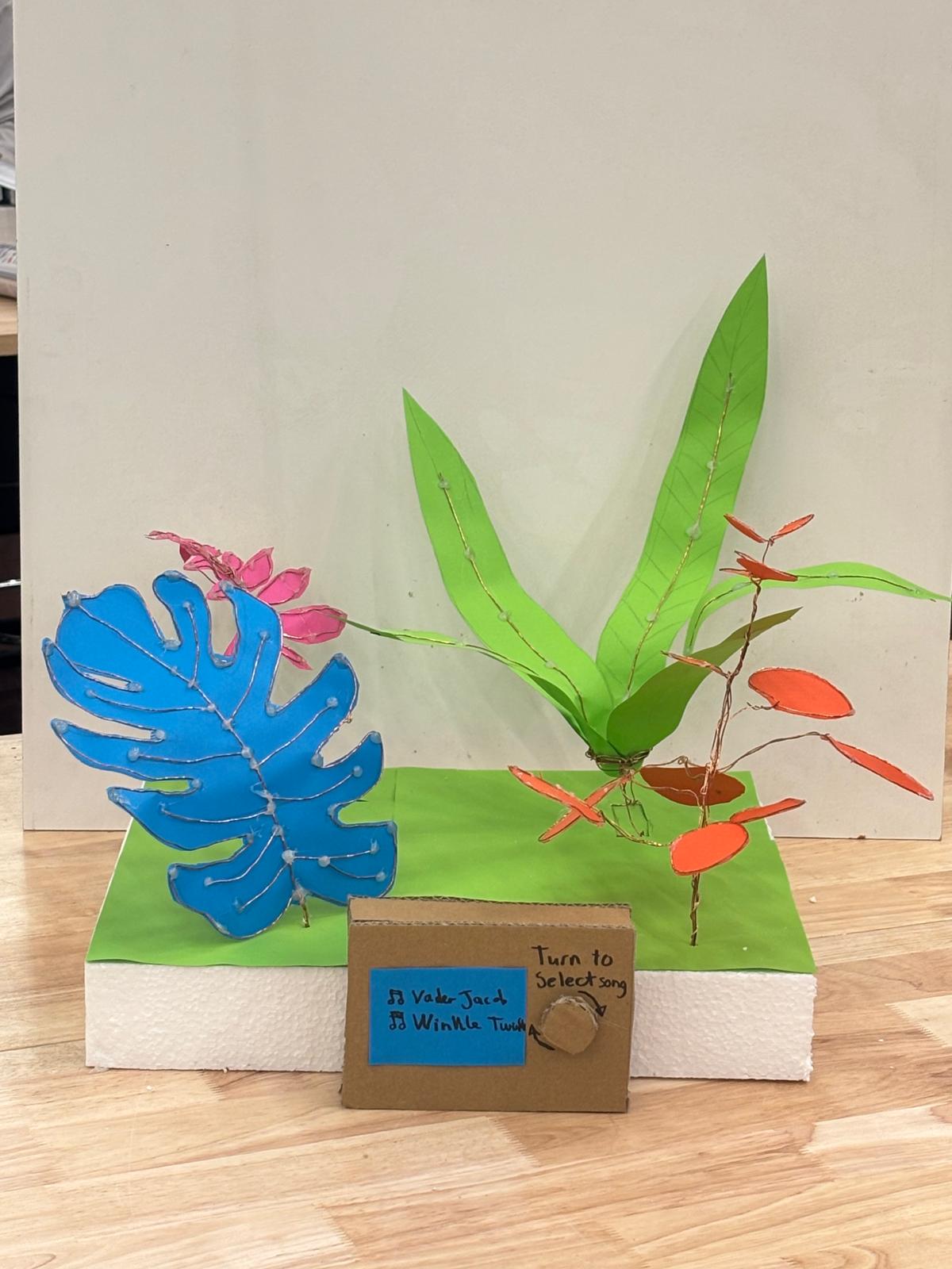 Early low-fidelity cardboard mockup: a paper leaf and plant next to a cardboard box labeled Turn to select song with two handwritten song names